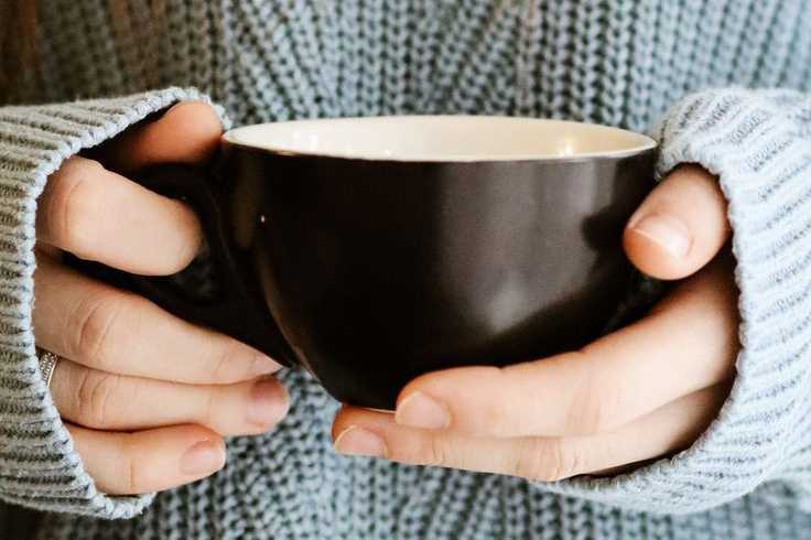 A woman holding a cup, wearing a blue sweater and have brunette hair.
