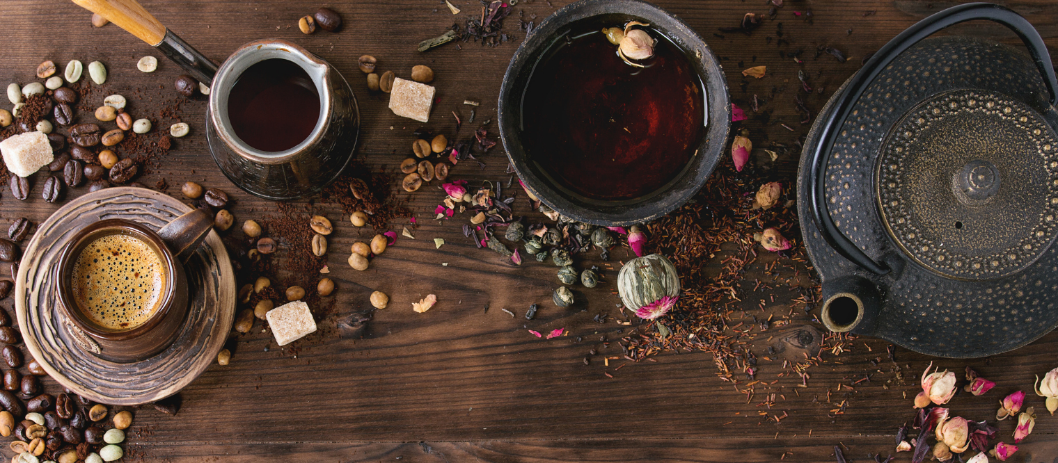 wooden background with black teacup vs coffee mug and black ancient teapot, coffee beans and dried tea leaves and rose petals all over the surface.