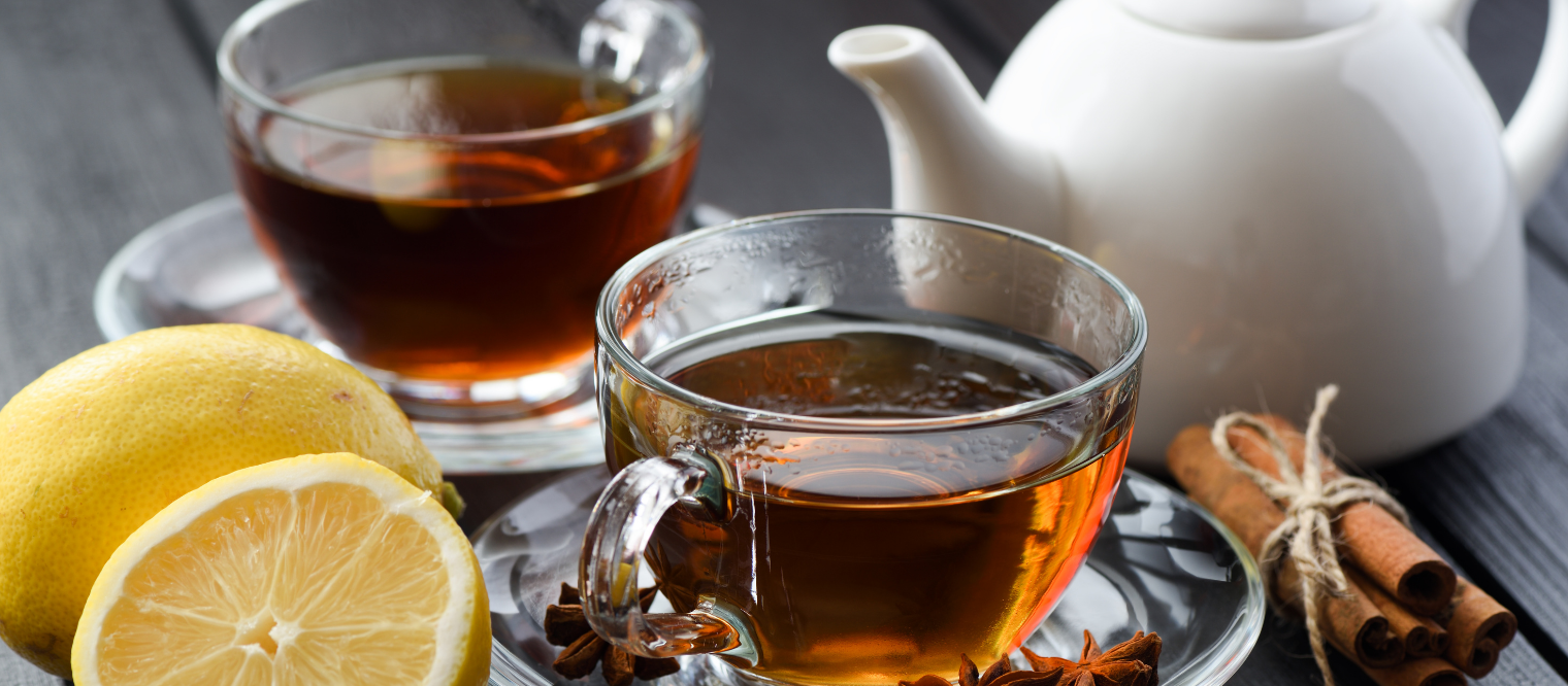 Black wooden background with white teapot and glass cup filled with black tea and lemon on side of the cup and cinnamon.