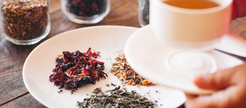 Tasting different types of tea, herbs and ingredients of tea on awhite late with glass jars filled with ingredients and a man hand with a cup of tea with wooden table.