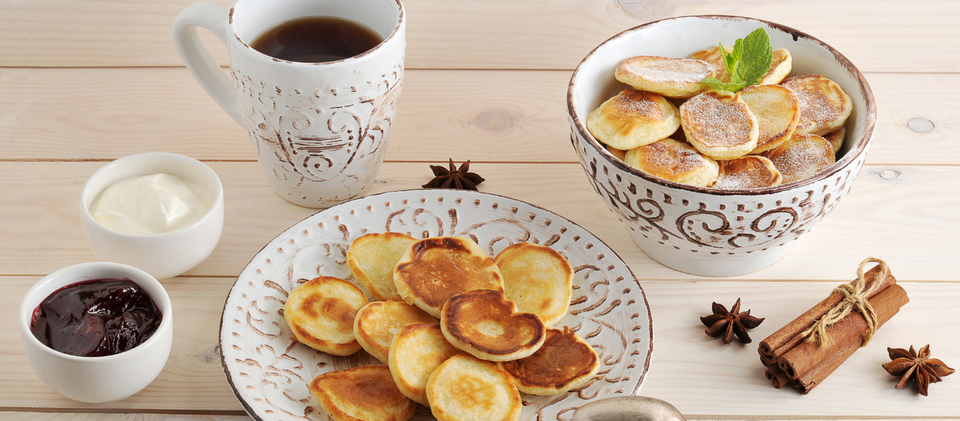 Traditional black tea in a white cup and crumpets on a white plate and in a white bowl, white cream, jam, and cinnamon on a wooden table.