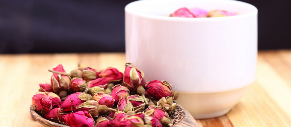 Rose petals on wooden spoon with wooden background and white tea cup filled with rose milk tea.